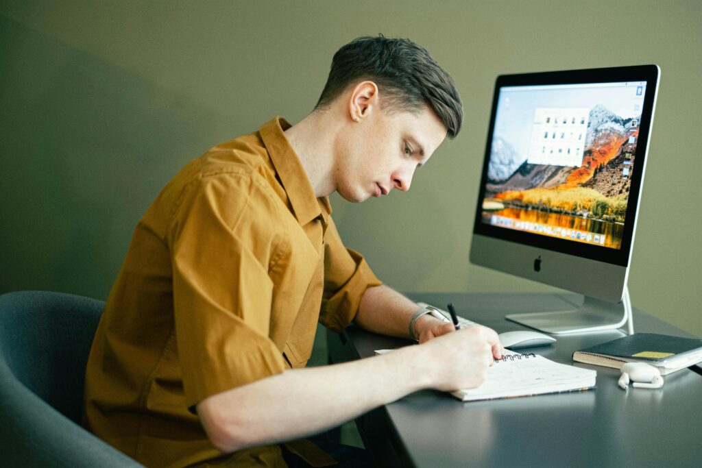 Adult male concentrating on notes with a desktop computer in a home office setting.