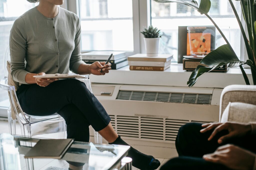 Unrecognizable female psychologist with crossed legs writing information on clipboard during appointment with anonymous ethnic patient in cozy light office