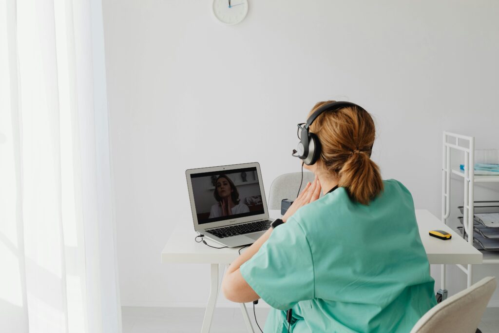A nurse in a teal uniform conducts a video call consultation from a modern clinic.